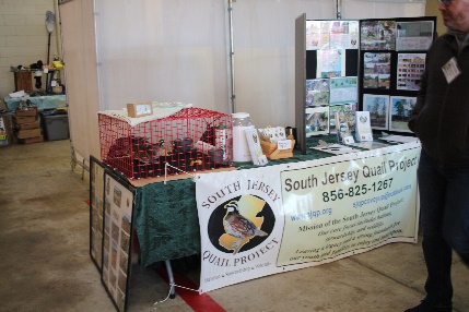 A cage with quail in it sit on top of a table with a SJQP banner across the front